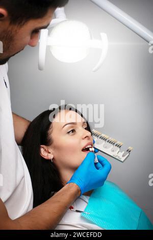 Eine Frau auf dem Zahnarztstuhl schaut auf seine Zähne und wählt die Farbe aus der Zahnpalette aus Stockfoto