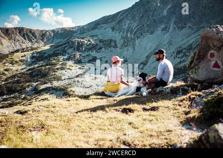 Ein Paar mit Hund sitzt auf Felsen und bewundert die Berglandschaft Stockfoto