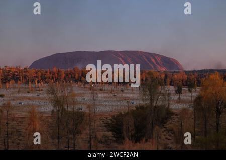 Letzte Sonnenstrahlen auf Uluru und die Field of Light Art Installation des Künstlers Bruce Munro. Flecken von Gras Feuer Rauch auf der Horizontlinie Stockfoto