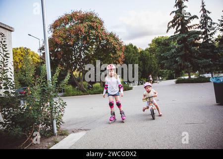 Zwei kleine Mädchen fahren auf Rollern und Laufrädern im Sommerpark. Kinder tragen Schutzpolster und Schutzhelm für eine sichere Fahrt. Aktiver Outdoor-Sport f Stockfoto