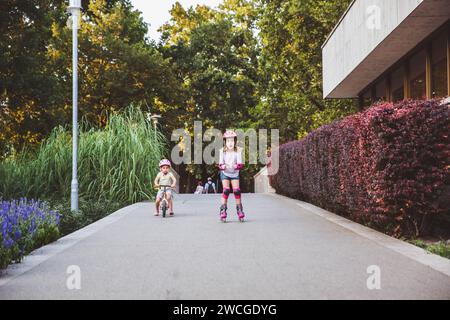 Zwei kleine Mädchen fahren auf Rollern und Laufrädern im Sommerpark. Kinder tragen Schutzpolster und Schutzhelm für eine sichere Fahrt. Aktiver Outdoor-Sport f Stockfoto