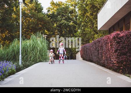 Zwei kleine Mädchen fahren auf Rollern und Laufrädern im Sommerpark. Kinder tragen Schutzpolster und Schutzhelm für eine sichere Fahrt. Aktiver Outdoor-Sport f Stockfoto