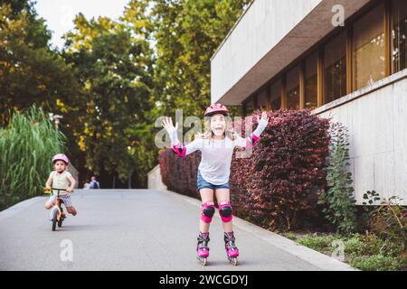 Zwei kleine Mädchen fahren auf Rollern und Laufrädern im Sommerpark. Kinder tragen Schutzpolster und Schutzhelm für eine sichere Fahrt. Aktiver Outdoor-Sport f Stockfoto