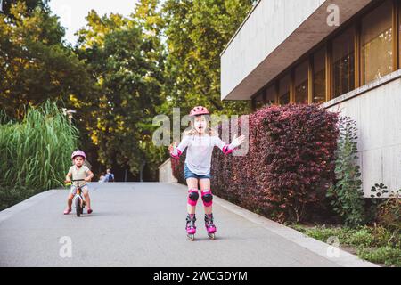 Zwei kleine Mädchen fahren auf Rollern und Laufrädern im Sommerpark. Kinder tragen Schutzpolster und Schutzhelm für eine sichere Fahrt. Aktiver Outdoor-Sport f Stockfoto