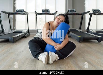 Eine Frau in Übergröße sitzt auf dem Boden und hält eine blaue Trainingsmatte und ruht sich während ihres Workouts aus. Stockfoto