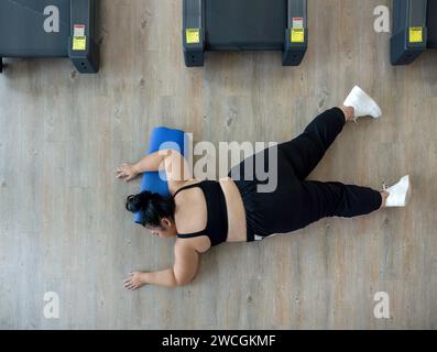 Eine Frau in Übergröße liegt auf dem Boden des Fitnessstudios, ruht oder unmotiviert, umgeben von Trainingsgeräten. Stockfoto