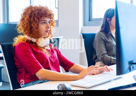 Rothaarige Frau mit Kopfhörern, die am Computer in einer hellen Büroumgebung arbeitet. Stockfoto