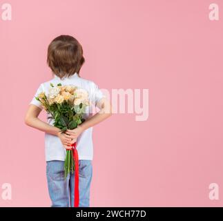 Ein kleiner Junge hält einen Rosenstrauß hinter dem Rücken auf rosa Hintergrund. Konzept von Feiertagen, Geburtstag, Valentinstag und Muttertag. Stockfoto