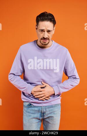 Bärtiger Mann in lila Pullover und Jeans, der an Magenschmerzen auf orangefarbenem Hintergrund leidet, Schmerzen Stockfoto