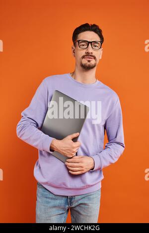 Bärtiger Mann in Brille und lila Pullover, der Laptop hält und auf orangefarbenem Hintergrund steht Stockfoto