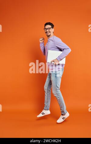 Glücklicher bärtiger Mann in Brille und lila Pullover, der mit Laptop auf orangem Hintergrund läuft Stockfoto