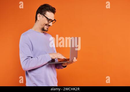 Lächelnder Mann in Brille und lila Pullover mit Laptop auf orangem Hintergrund, Fernarbeit Stockfoto