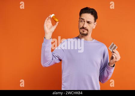 Bärtiger Mann im lila Sweatshirt, der Flasche mit Pillen und Blisterpackung auf orangem Hintergrund hält Stockfoto