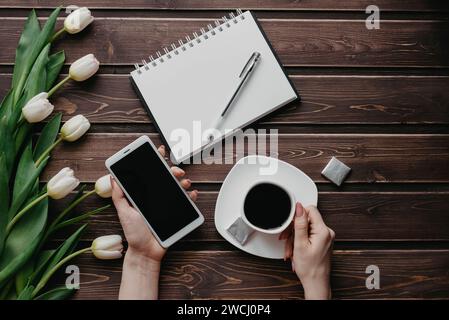 Weiße Tulpen auf einem Holztisch mit leerem Notebook, Smartphone und einer Tasse Kaffee in Frauenhänden. Flache Anordnung, Draufsicht mit Kopierraum Stockfoto