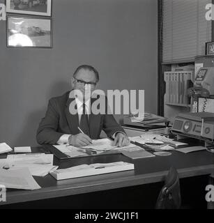 1970er Jahre, historisch, ein Geschäftsmann im Automobilhandel, an seinem Schreibtisch mit Papieren und Stift in der Hand, ein Autohändler, möglicherweise ein Buchhalter, wie auf dem Schreibtisch eine Broschüre über nationale Versicherungsbeiträge. Auf der rechten Seite ein Telefonsystem der Ära, mit einem Telefon, das auf einem Ständer mit Wählschalter unten hängt. Stockfoto