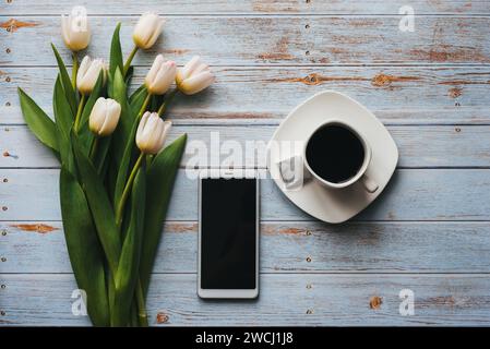 Weißer Tulpenstrauß auf blauem hölzernem Hintergrund mit Tasse Kaffee und einem Smartphone. Flache Lage, Draufsicht. Leere Grußkarte für Valenti Stockfoto