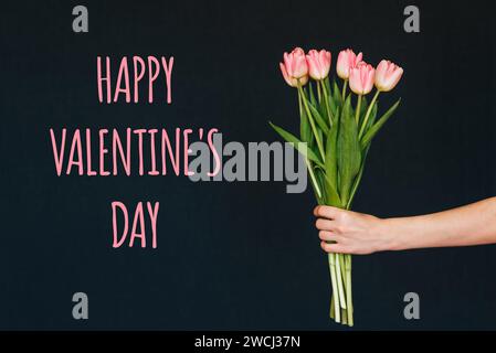 Grußkarte mit der Inschrift Happy valentine's Day. Strauß rosa Tulpenblüten in der Hand einer Frau auf einem Hintergrund mit Glückwunschtext Stockfoto