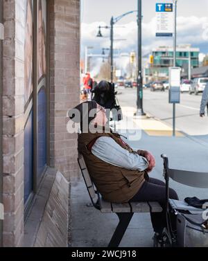 Ein älterer Mann in warmer Kleidung sitzt an einer Bushaltestelle. Ein älterer kaukasischer Mann sitzt auf der Bank und wartet auf den Bus. Der alte Rentner wartet auf Transport-Vanc Stockfoto