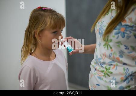 Mutter sprüht Nasenspray gegen das Fieber ihrer Tochter, das kleine Mädchen wird zu Hause behandelt und ist im Pyjama - verschwommener Hintergrund. Stockfoto