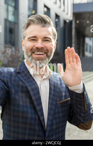 Ein lächelnder reifer Geschäftsmann in einem stilvollen Anzug winkt Hallo, steht draußen an einem Bürogebäude und strahlt Führungsstärke aus. Stockfoto