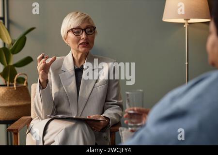 Porträt einer reiferen Therapeutin, die eine Brille trägt und mit nicht erkennbarem Klienten spricht, während einer Sitzung, Kopierraum Stockfoto