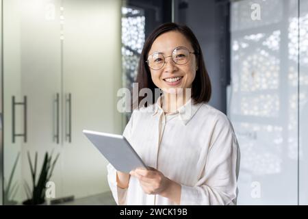 Professionelle junge asiatische Geschäftsfrau mit einem Tablet, die selbstbewusst an ihrem Schreibtisch in einem modernen Büro lächelt. Stockfoto