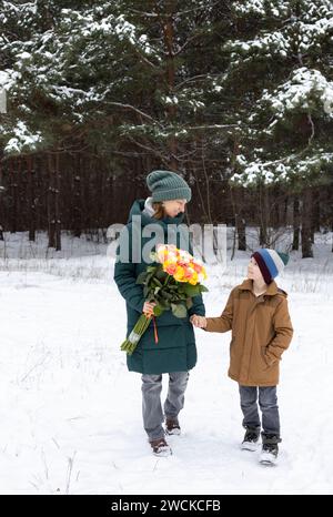 Mom hält ihren Sohn mit einer Hand und die andere Hand hält einen großen Rosenstrauß. Eine Frau und ein Kind laufen im Winter zusammen im Park in A Stockfoto