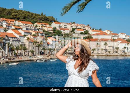 Eine elegante Frau steht anmutig am ruhigen Wasser und strahlt zeitlose Schönheit in ihrem fließenden weißen Kleid und ihrem stilvollen Strohhut aus Stockfoto