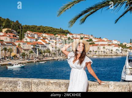 Eine atemberaubende junge Frau in einem eleganten weißen Kleid steht anmutig am malerischen Meer, mit einer bezaubernden Altstadt als Hintergrund Stockfoto