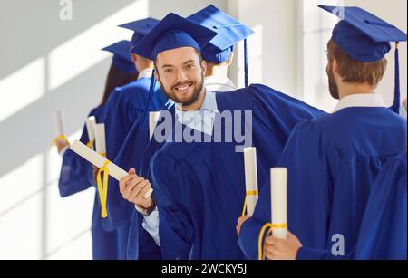 Die Abschlussfeier der Studenten, glückliche Erwachsene Studenten in Reihe, die eine komplette Outfit-Mütze und ein blaues Kleid tragen Stockfoto