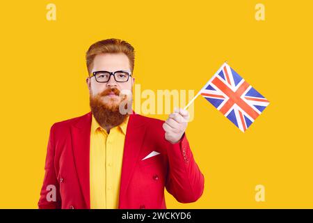 Stilvoller bärtiger Mann mit ernstem und selbstbewusstem Ausdruck, der die britische Flagge auf gelbem Hintergrund hält. Stockfoto