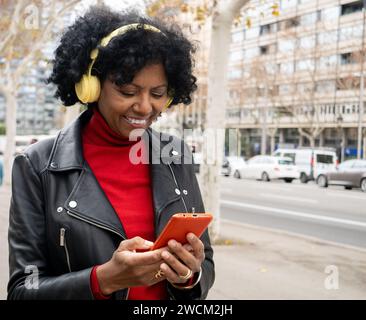 Sorglose Afroamerikanerin mittleren Alters, die in einer Stadt mit Smartphone und Kopfhörern chatten Stockfoto