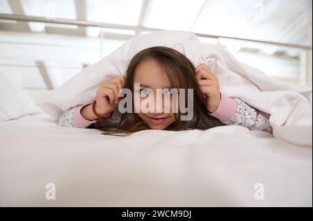 Weißes entzückendes glückliches, schelmiges kleines Mädchen in stilvollem Pyjama, versteckt sich unter weißer Decke, während sie auf ihrem Bett in der gemütlichen Schlafkammer zu Hause liegt Stockfoto