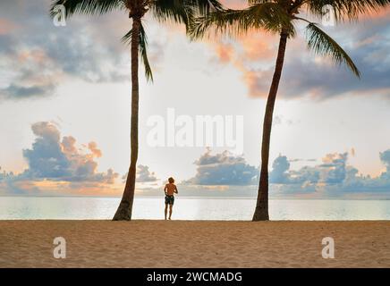 Ein laufender Teenager zwischen hohen Palmen am Strand von Flic en Flac mit einem unglaublichen Sonnenuntergangshimmel. Insel Mauritius. Exotische Reise- und Urlaubshindernisse Stockfoto