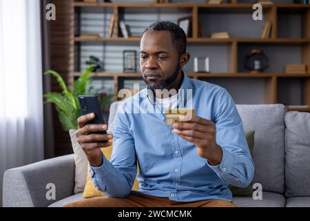 Besorgter Afroamerikaner, der auf das Handy schaut. Auf der Couch sitzen und eine Kreditkarte halten. Stockfoto