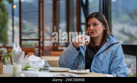 Schöne junge lateinamerikanische Frau, die in einem Restaurant sitzt, eine Tasse Kaffee in der Hand, eine blaue Jacke trägt, mit einer nachdenklichen Haltung Stockfoto