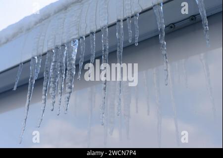 Eiszapfen hängen von einer Dachrinne an einem klaren Himmel mit Schnee am Rand und Reflexion auf glänzenden Sockeln. Stockfoto