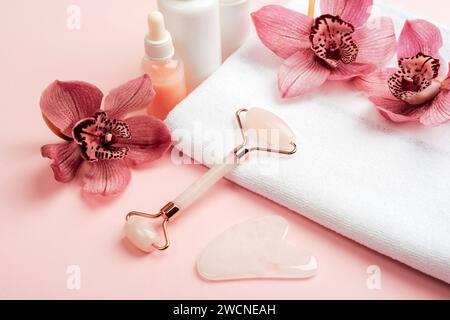 Kosmetikrolle und Gua-Sha-Schaber mit rosa Orchideenblüten auf einem weißen Handtuch. Home Spa Konzept. Stockfoto
