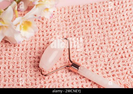 Gesichtsrolle aus Rosenquarzkristall auf rosa Handtuch und weißer Orchideenblüte. Gesichtsmassagegerät zur Lifting-Therapie. Draufsicht, flach. Stockfoto