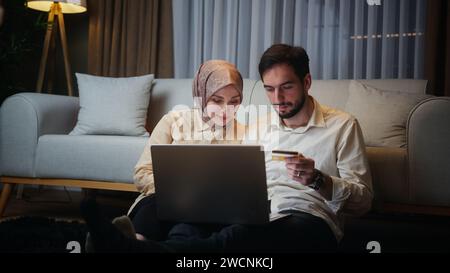 Junge verheiratete Paare, die nachts auf dem Boden sitzen und mit einem Laptop online einkaufen, mit einer Kreditkarte im Besitz des Mannes Stockfoto