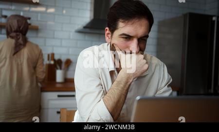 Seriöser Ehemann, der online budgetiert, bezahlt Rechnungen und Finanzen, Hypothekenzahlungen mit Laptop-Computer, während seine Frau in der Küche arbeitet Stockfoto