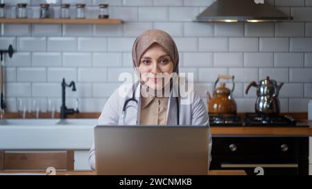 Die Ärztin im Kopftuch trägt einen weißen Mantel und spricht mit der Kamera, während sie in der Küche sitzt Stockfoto