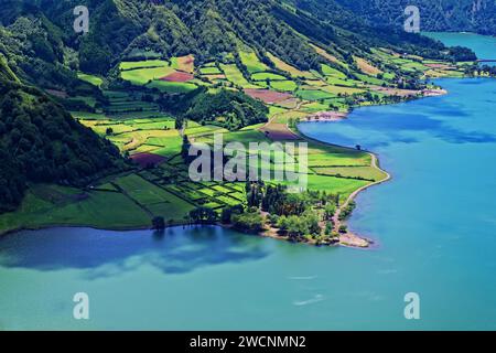 Blick auf die Kulturlandschaft mit grünen Feldern neben dem blauen Wasser des Kratersees Lagoa Azul, Lagoa Azul, Rundwanderweg, Caldeira Stockfoto