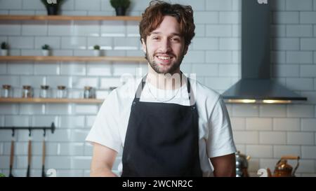 Porträt eines jungen Mannes mit lockigen Haaren mit Schürze, der in die Kamera blickt und in der Küche lächelt Stockfoto