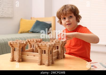 Süßer kleiner Junge, der mit einer hölzernen Festung am Tisch im Zimmer spielt. Kinderspielzeug Stockfoto