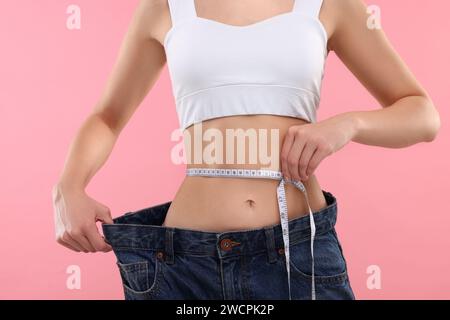 Schlanke Frau, die große Jeans trägt und die Taille misst, mit Klebeband auf rosa Hintergrund, Nahaufnahme. Gewichtsverlust Stockfoto