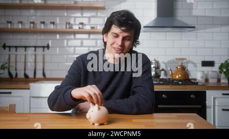 Ein glücklicher Mann, der zu Hause in einer modernen Küche sitzt, steckt ein paar Münzen in die Schweinebank Stockfoto