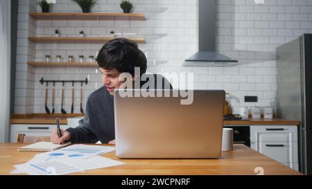 Ein lächelnder junger Student sitzt zu Hause in der Küche, sieht nach einem Laptop, lernt, macht Notizen. Männlicher Student, der recherchiert, studiert an der h Stockfoto