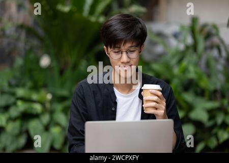 Ein positiver junger asiatischer Freelancer oder digitaler Nomad, der aus der Ferne in einem Café arbeitet, Kaffee trinkt und an seinem Laptop arbeitet. Urbanes Zitat Stockfoto