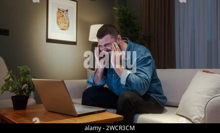 Junger erwachsener Mann, der zu Hause auf dem Sofa sitzt, während er einen Laptop benutzt und Kopfschmerzen hat. Müde junge Männer bekommen Schmerzen durch Kopfschmerzen oder Migräne Stockfoto